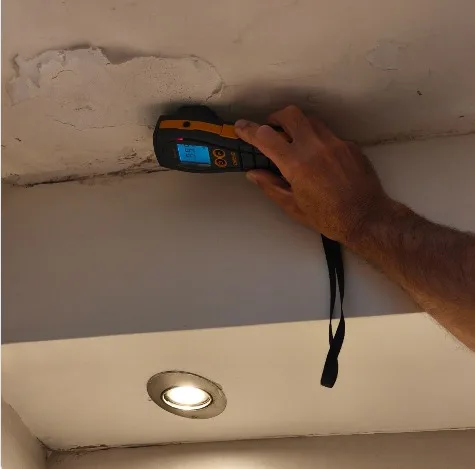 מומחה בדק בית מצמיד מד לחות לתקרת בטון מתקלפת מעל תקרת גבס מונמכת עם גוף תאורה שקוע - איתור רטיבות נסתרת וחשד לעובש