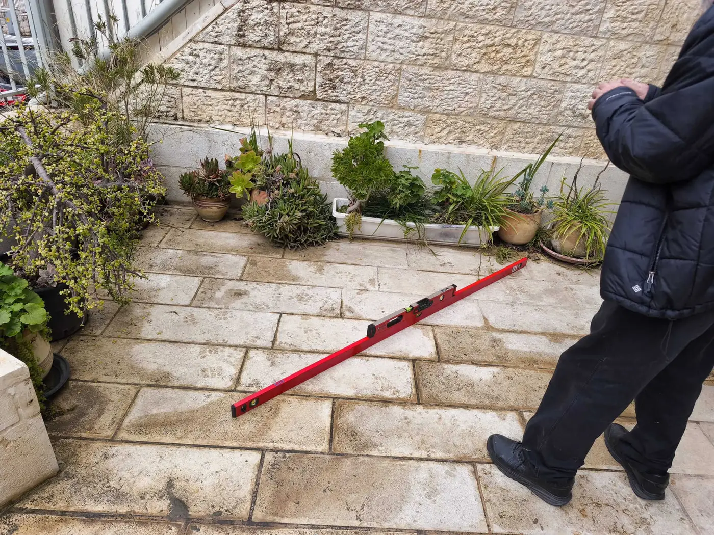 מהנדס בדק בית מודד את שיפוע הריצוף בחצר בית פרטי בעזרת פלס דיגיטלי ארוך המונח בניצב לקיר האבן - בדיקת ניקוז והרחקת מים מהמבנה